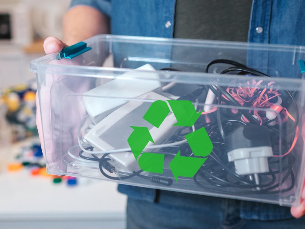 Caja de plástico transparente con símbolo de reciclaje que contiene multitud de cables y aparatos electrónicos sujetado por un chico