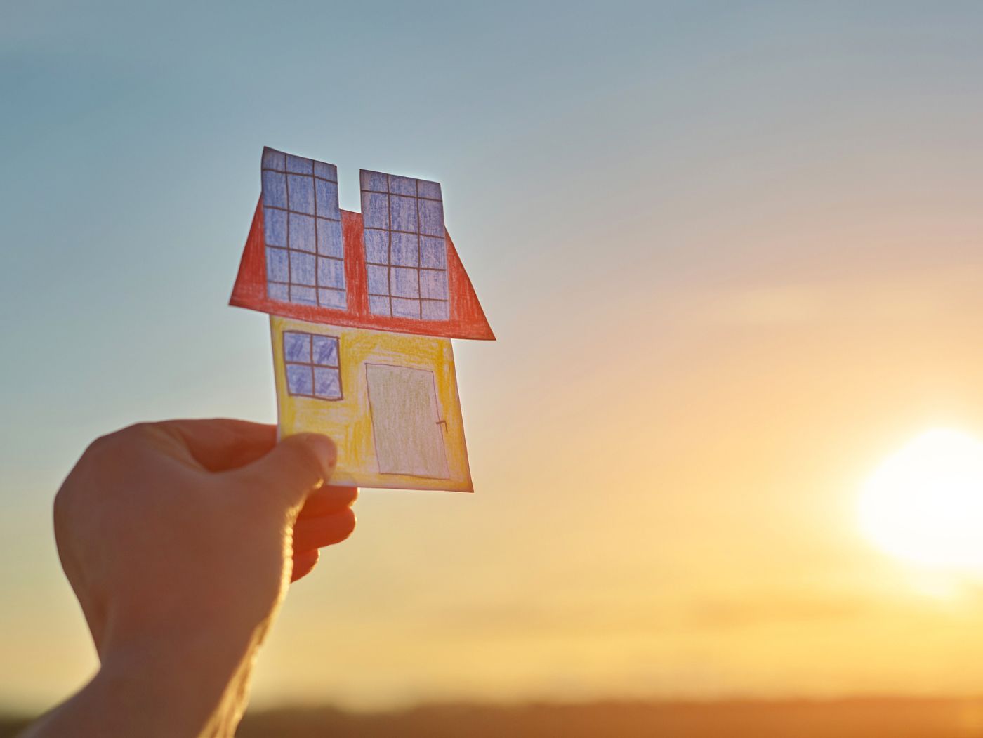 Imagen de una mano sujetando un dibujo de una casa con energía solar en el techo, De fondo el sol atardeciendo o amaneciendo