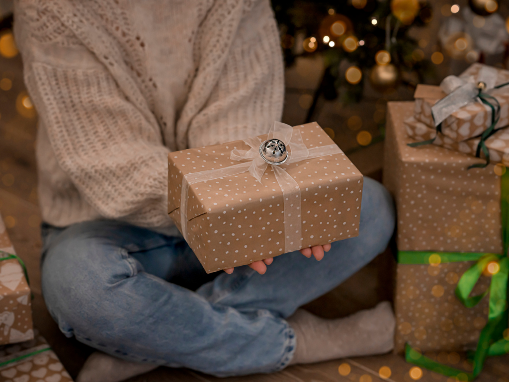 Una persona sentada en el suelo junto a un árbol de navidad y regalos sujeta un regalo
