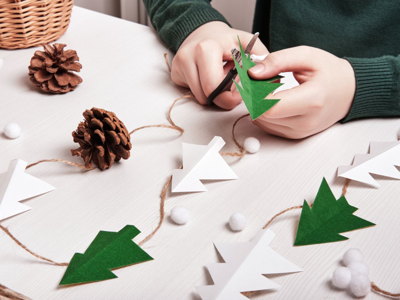 Plano de unas manos recortando cartulinas con forma de árbol de Navidad. En la mesa hay árboles de cartulina conectados por una cuerda y varias piñas sobre la mesa